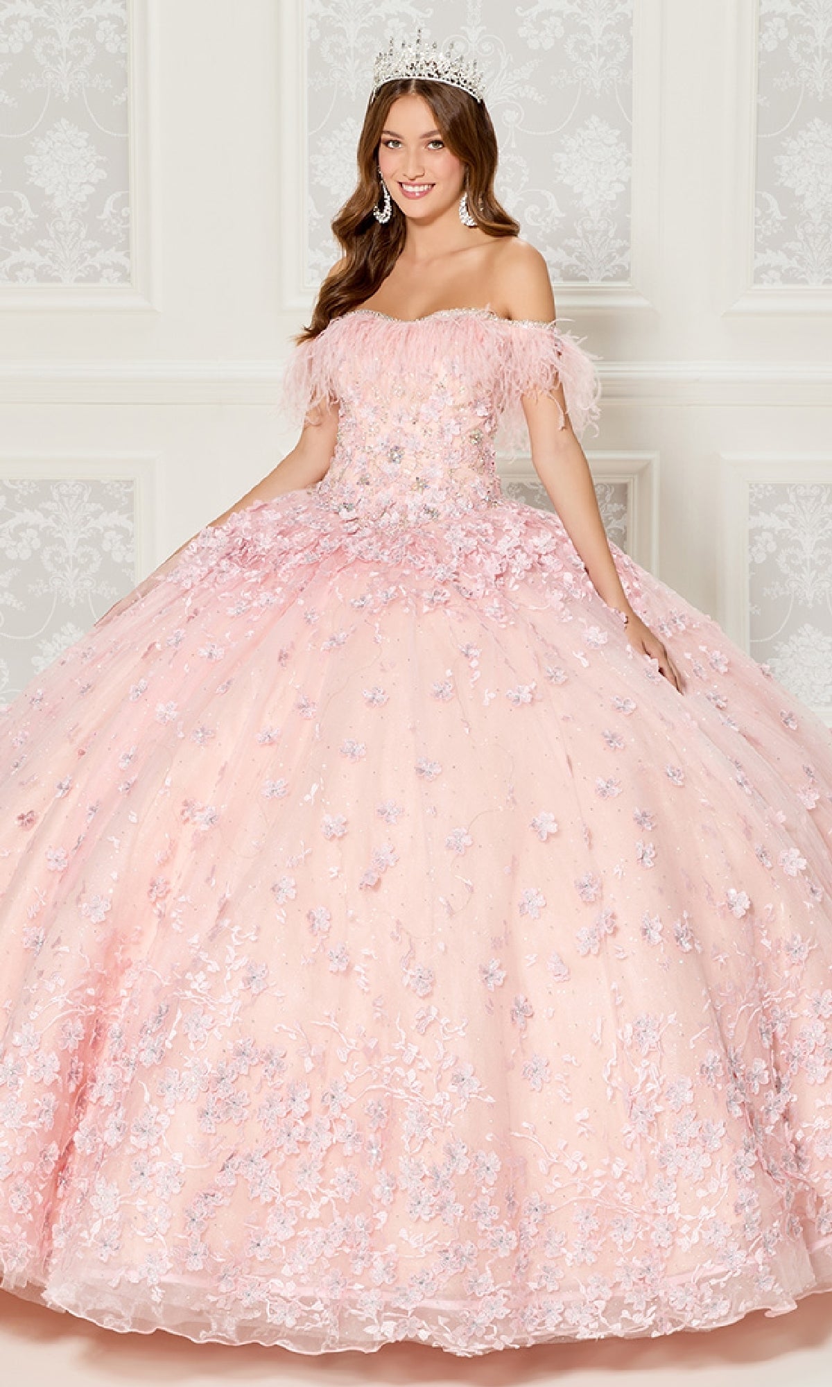 A young woman in the Quinceanera Dress PR30115 By Princesa, featuring pink off-the-shoulder floral details and a tiara, stands in an elegant, white-paneled room.